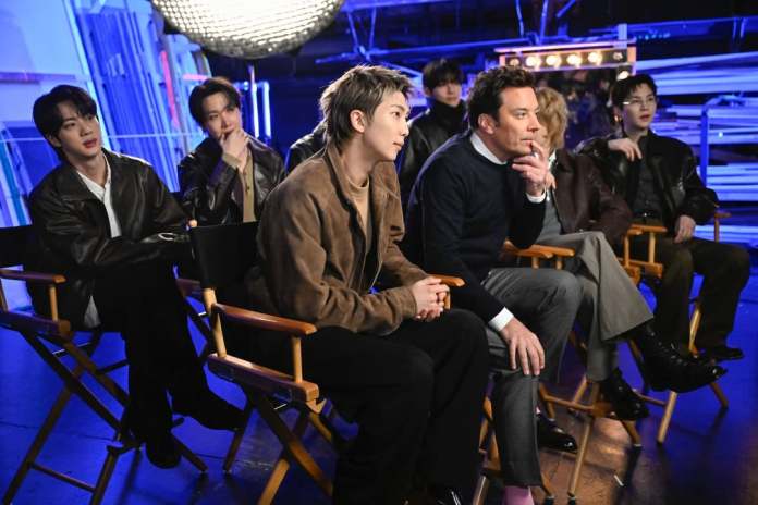 BTS members and Jimmy Fallon sitting in director's chairs under studio lights, with one person in the foreground looking thoughtful. The setting appears to be a television studio.