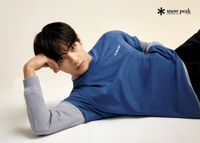 V with dark hair poses casually while lying on a light background, wearing a blue and gray long-sleeve shirt with the logo 'snow peak.'