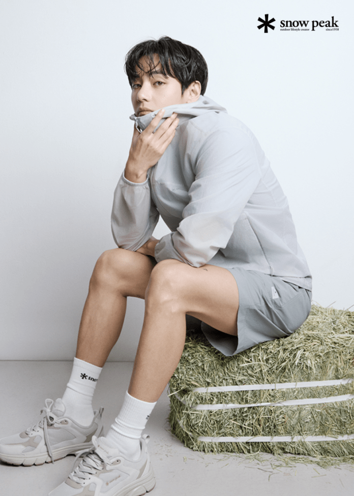 V wearing a light gray jacket and shorts sits on a bale of hay, thoughtfully gazing at the camera. He has dark, wavy hair and is accessorized with light sneakers and white socks.