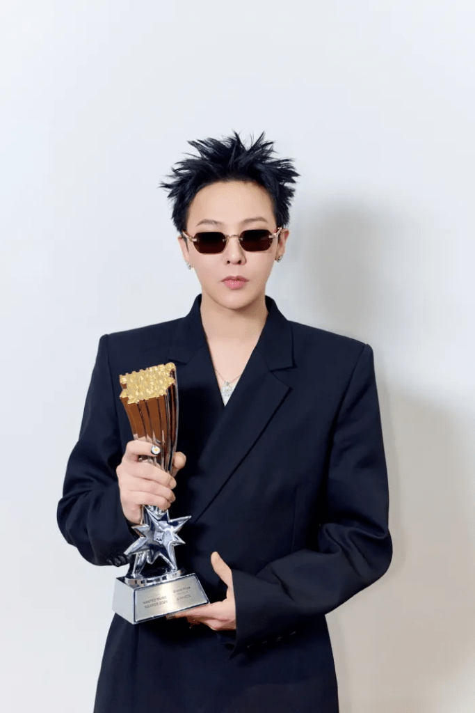GD wearing sunglasses and a black oversized suit holding a trophy with a star base, posing against a plain white background.
