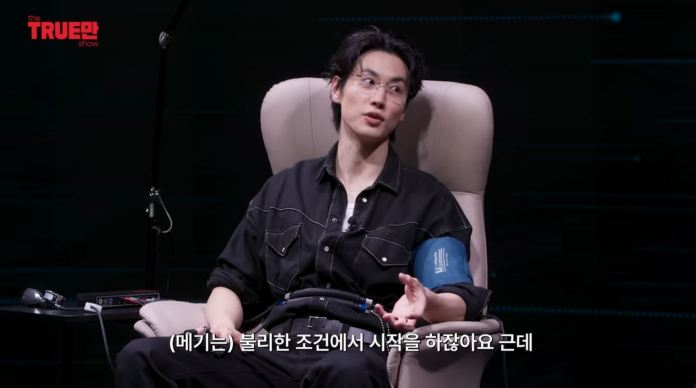 Jo igeon sitting in a chair, wearing a black shirt, discussing while connected to a medical monitoring device on their arm, with a softly lit background.