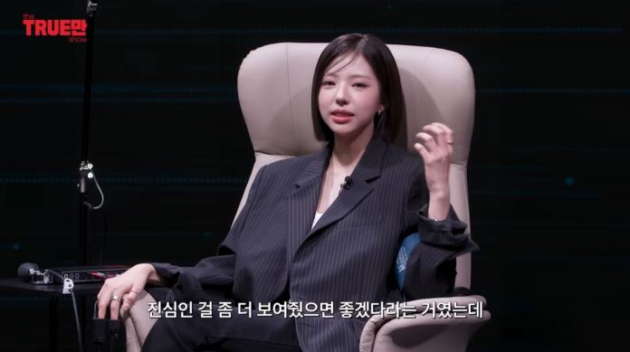 Kim Go Eun in a striped blazer sits comfortably in a chair, gesturing as she speaks. The background features a digital design, and there is text in an Asian language at the bottom.