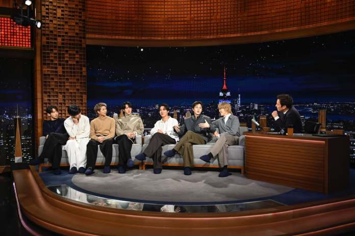 BTS members and Jimmy Fallon sitting on a talk show stage set, engaging in conversation. The backdrop features a city skyline with a visible landmark. Jimmy Fallon is seated at a desk, listening intently to BTS members.