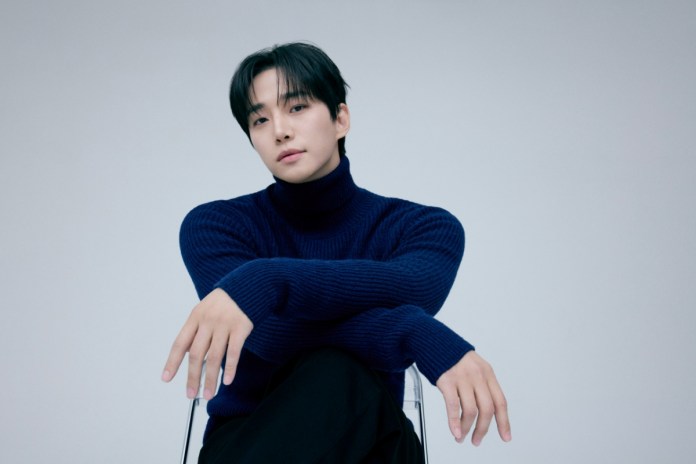 Lee Junho with dark hair wearing a blue turtleneck sweater, seated in a transparent chair, with his arms crossed and a neutral expression against a light gray background.