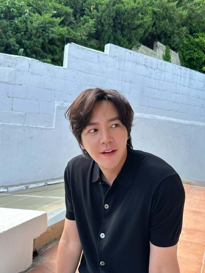 Jang Keun-suk with dark, wavy hair wearing a black polo shirt, sitting outdoors against a backdrop of greenery and a white wall.