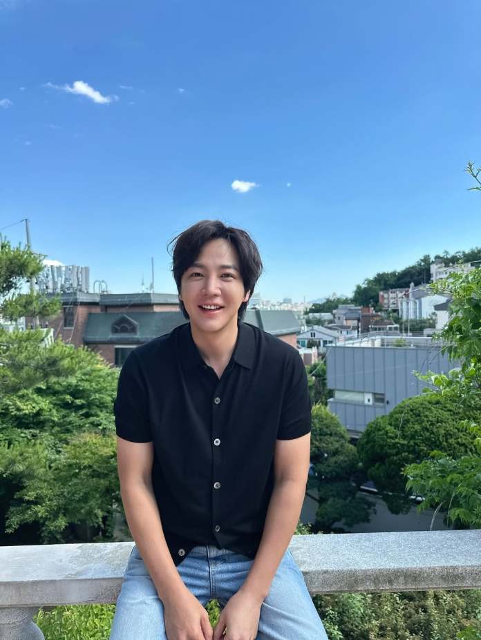 Jang Keun-suk with short black hair smiles while sitting on a railing, wearing a black button-up shirt and blue jeans, with a green landscape and blue sky in the background.