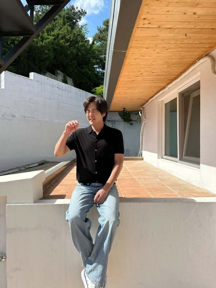 Jang Keun-suk sitting on a ledge in casual clothing, smiling and making a hand gesture, with a sunny outdoor setting featuring a modern house and greenery in the background.