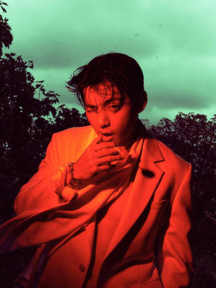 V in a stylish light-colored suit poses dramatically, holding his finger to his lips in a suggestive manner. The image is illuminated with warm red tones against a greenish background with foliage.