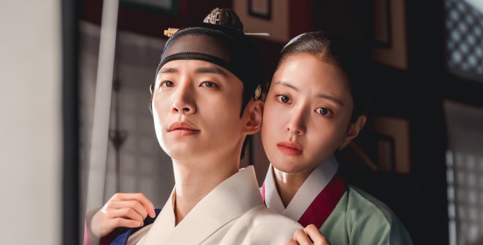 Lee Junho in traditional Korean attire, with the woman gently embracing the man from behind, showcasing an intimate moment in a historical setting.