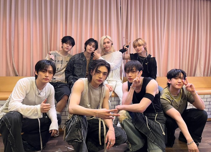 Stray Kids posing together, showcasing a stylish and casual fashion aesthetic. They are smiling and using hand gestures, with a background of curtains and a wooden surface.