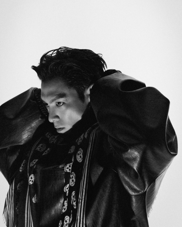 A close-up black and white portrait of T.O.P with slicked-back hair, wearing a leather jacket and a patterned scarf, gazing thoughtfully while adjusting their hair.
