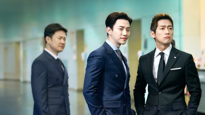 Lee Junho in formal suits standing in a modern office setting, with two men looking serious and the third man appearing contemplative in the background.
