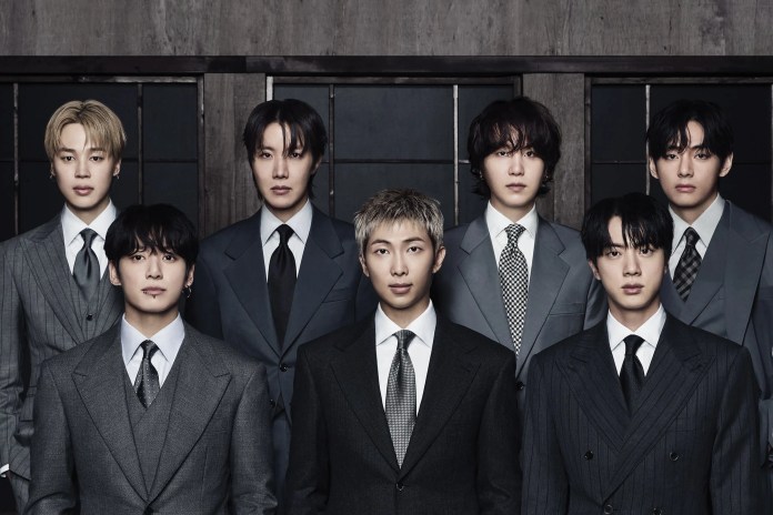 BTS individuals wearing formal suits, standing in two rows against a dark background. They display varied hairstyles and facial expressions, emphasizing a stylish, professional look.