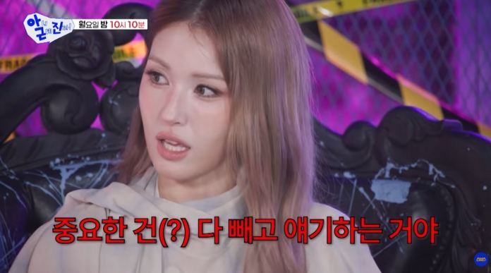 Jeon Somi with long hair appears surprised while speaking, with a decorative background featuring a black chair and colorful lighting.