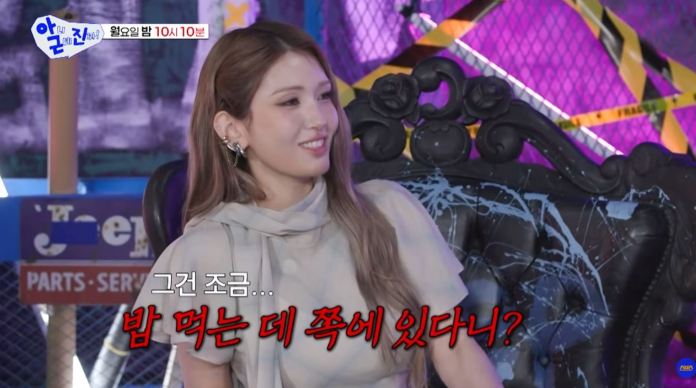 Jeon Somi with long hair, wearing a light-colored outfit, smiles while seated in an ornate black chair. The background features colorful decorations and caution tape, suggesting a fun or playful setting.