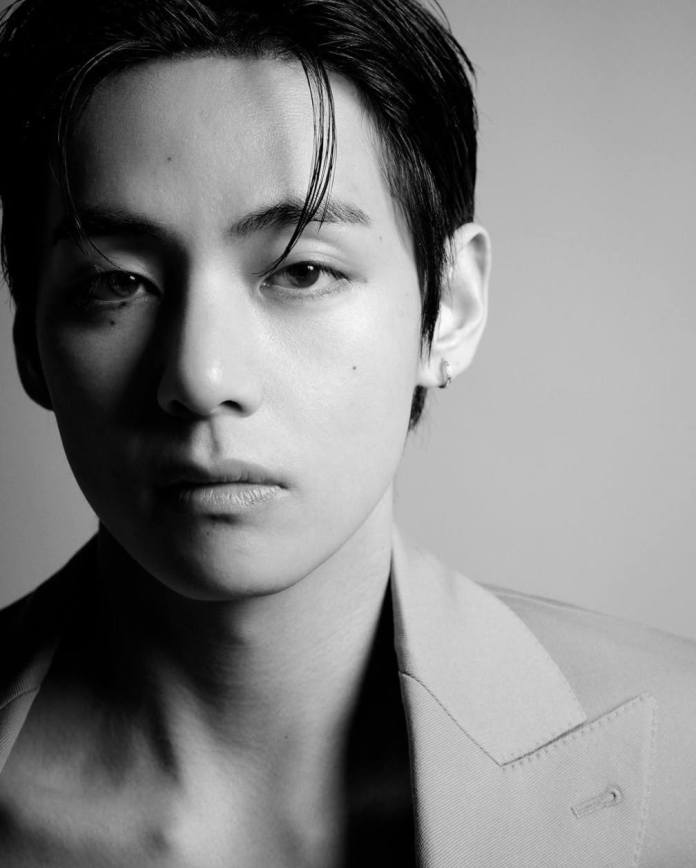 V in black and white, showing a serious expression with sharp features, styled hair, and a light-colored suit jacket.