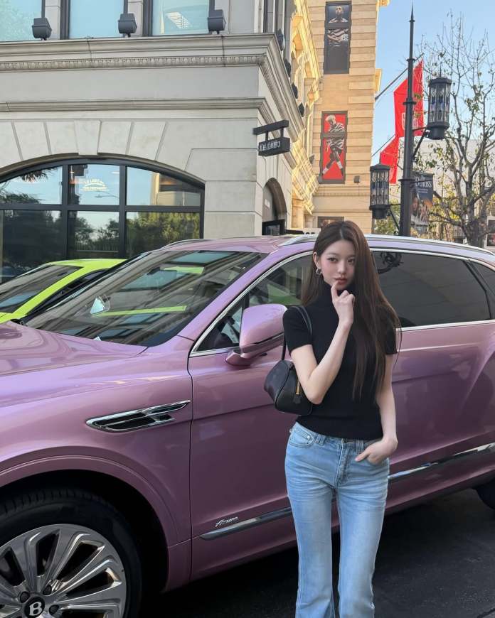 Jang Wonyoung in a black top and light blue jeans stands next to a pink luxury SUV, posing thoughtfully with her finger near her chin. The background features a stylish building and banners, indicating a lively urban setting.