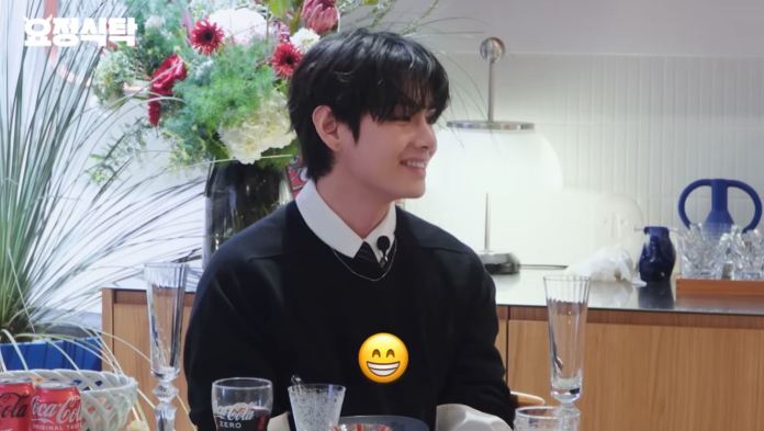 BTS V with dark, wavy hair is smiling while seated at a table adorned with flowers and various drinkware.