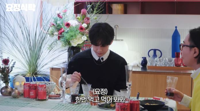 BTS V sitting at a table with food and drinks, looking down as he prepares to eat, with a colorful background featuring plants and decorative items.