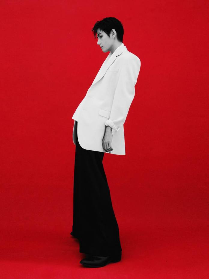 BTS’s V wearing a white blazer and black pants, standing against a bright red background.