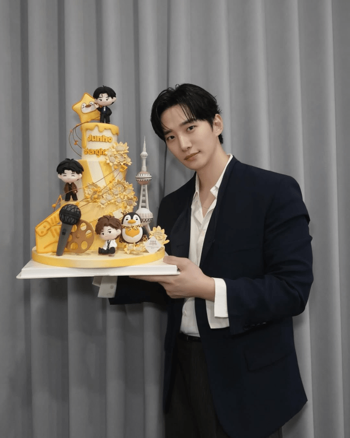 Lee Junho holding a colorful birthday cake decorated with figurines and themed elements, including a microphone and a penguin, set against a backdrop of gray curtains.
