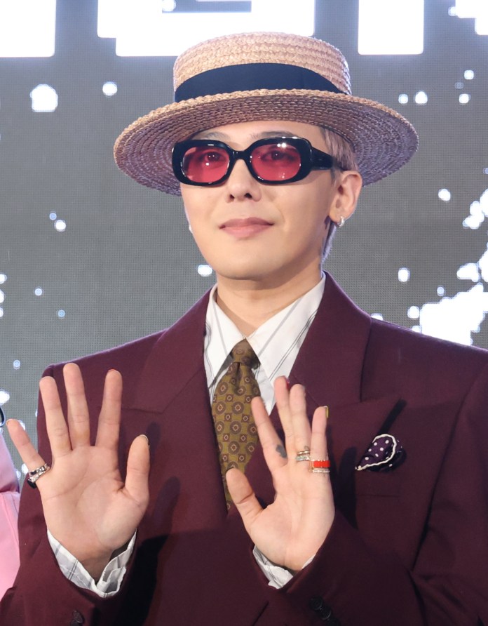 G-Dragon wearing a straw hat, oversized dark sunglasses, and a burgundy suit, posing with hands raised in a welcoming gesture.