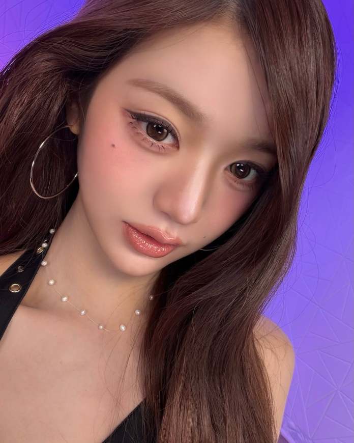 Close-up portrait of Jang Wonyoung with long, straight brown hair, wearing a black outfit and pearl necklace, against a purple background.