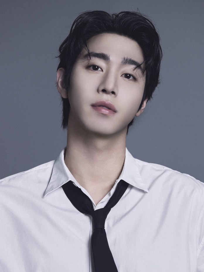 A young man with dark hair and a slight smile, wearing a white collared shirt and a black tie, against a gray background.