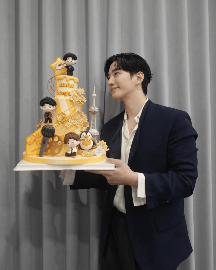 Lee Junho in a suit holding a colorful birthday cake decorated with figurines, including music and film themes, in front of a gray curtain.