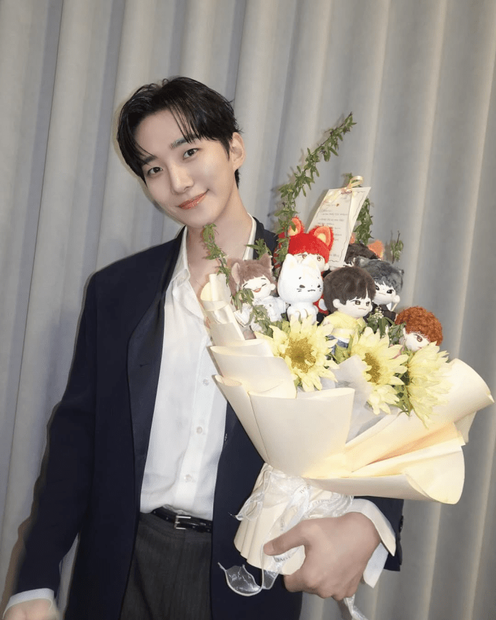 Lee Junho holding a large bouquet featuring sunflowers and plush doll characters, wearing a black blazer over a white shirt.