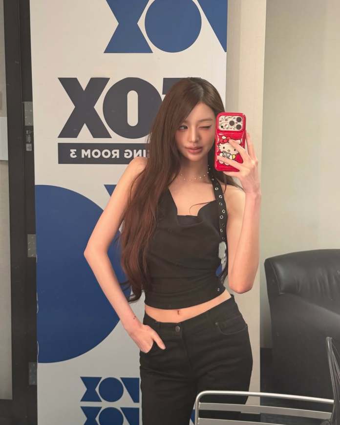 Jang Wonyoung taking a selfie in front of a mirror, wearing a black sleeveless top and black pants, with long hair and a wink. The background features a blue and white logo design.