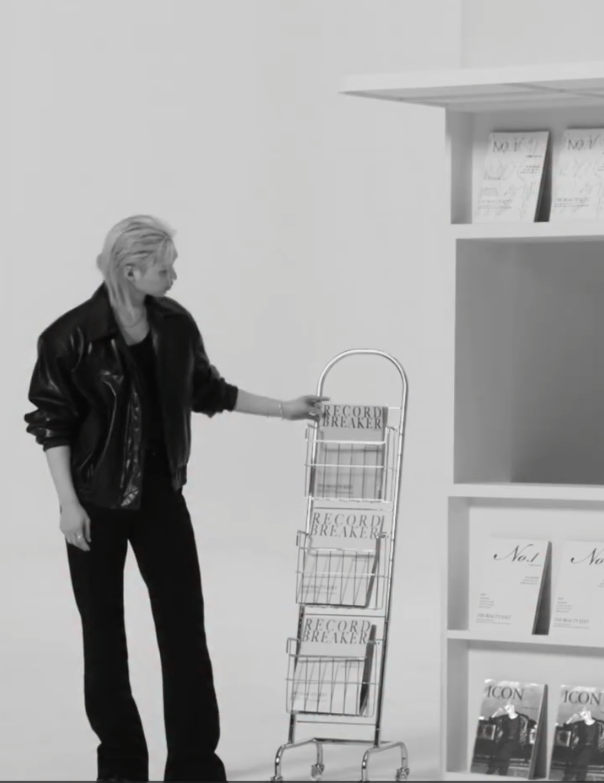Felix in a black leather jacket stands next to a metallic magazine rack displaying issues titled 'Record Breaker' and a bookshelf with various magazines in a minimalistic setting.