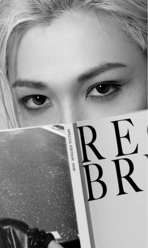 Portrait of Felix with platinum hair holding a magazine partially covering their face, displaying intense eyes and dramatic makeup, in black and white.