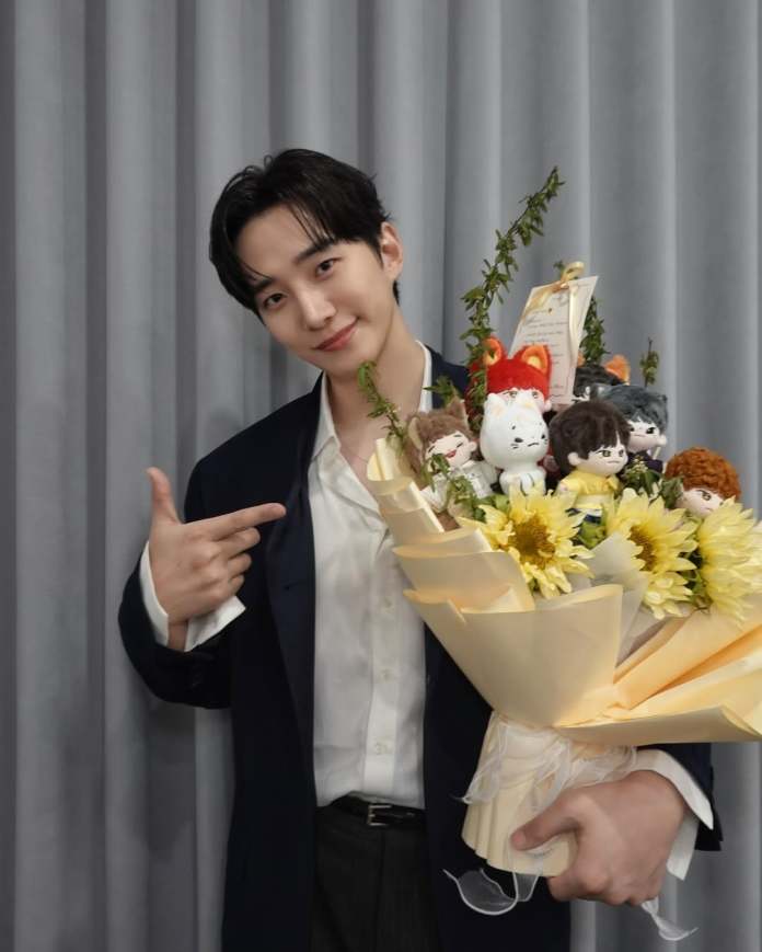 Lee Junho in a black jacket and white shirt poses with a large bouquet featuring sunflowers and plush toys, smiling and pointing.