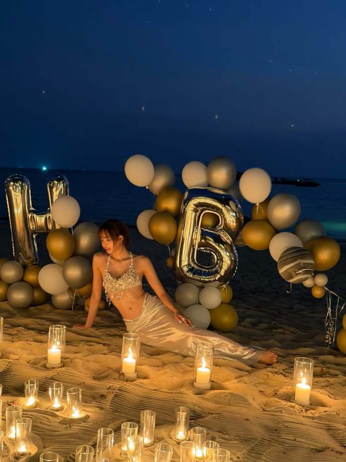 Lisa in a sparkling outfit poses on a beach at night, surrounded by candles and decorated with balloons spelling 'HB'.