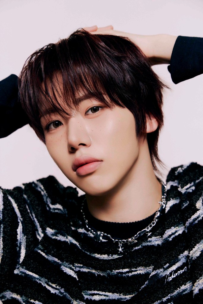 AHOF member with dark hair styled casually, wearing a striped sweater and a chain necklace, posing with hands behind their head against a soft, light background.