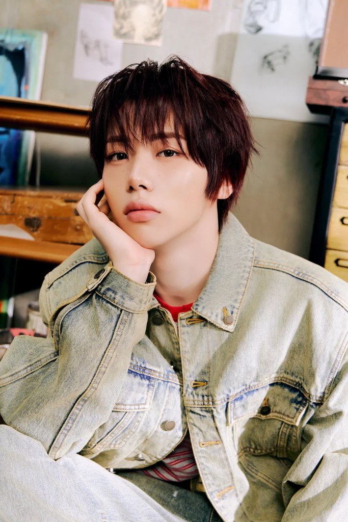 AHOF member posing with a thoughtful expression, wearing a denim jacket over a red shirt, with a studio background featuring artwork.