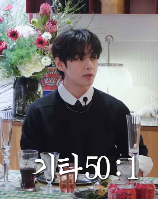 V wearing a black sweater and white collar sits at a table adorned with flowers, drinks, and dinnerware, looking thoughtfully.