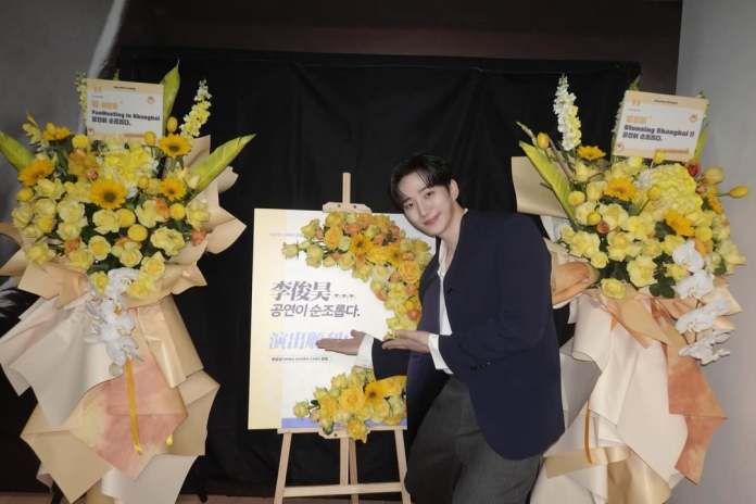 Lee Junho smiling next to a floral display featuring yellow flowers, standing beside a poster that includes the text 'Painting in Shanghai'.