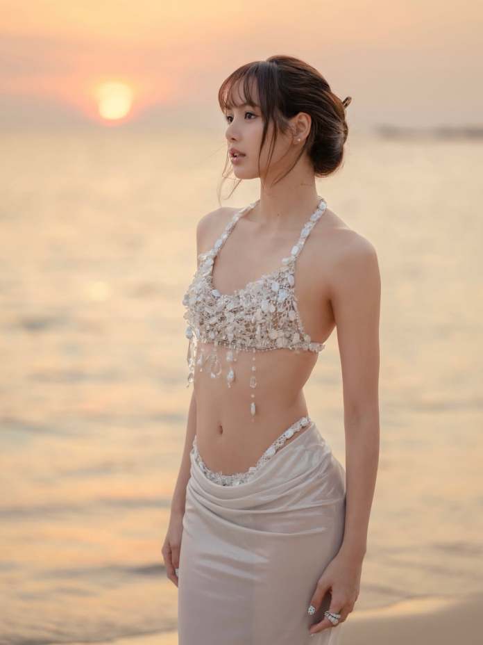Lisa wearing a beaded halter top and a sleek skirt stands by the beach during sunset, with the sun setting over the water in the background.