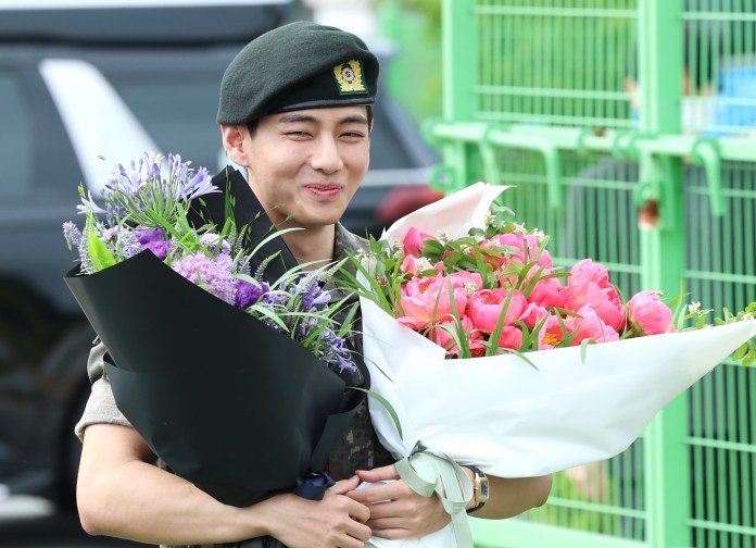 V in a military uniform and beret happily holds two large bouquets of flowers, one with pink peonies and the other with purple blooms.