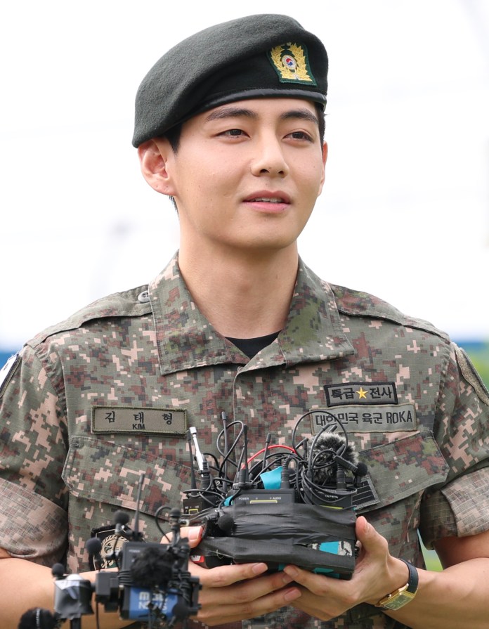 V in camouflage uniform and green beret, holding a bundle of audio and video recording equipment, with a focused expression.