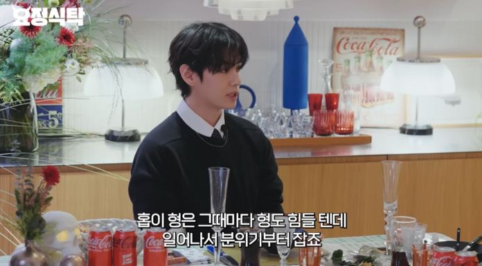 V with dark hair and a black outfit sitting at a dining table, engaged in conversation with decorative elements and various drinks visible.
