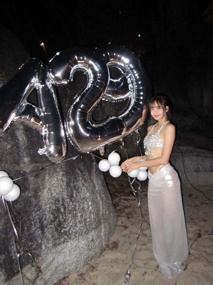 Lisa in a sparkling outfit stands beside large silver balloons that read '420' while holding a cluster of white balloons, set against a rocky and sandy background.