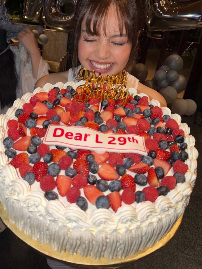 Lisa smiling joyfully while holding a decorated birthday cake topped with strawberries and blueberries, featuring a 'Happy Birthday' sign and a label that reads 'Dear L 29th'.