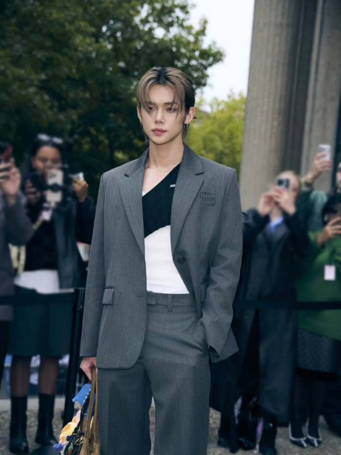 Yeonjun in a gray suit with a black shirt stands confidently outdoors, surrounded by people taking photos.