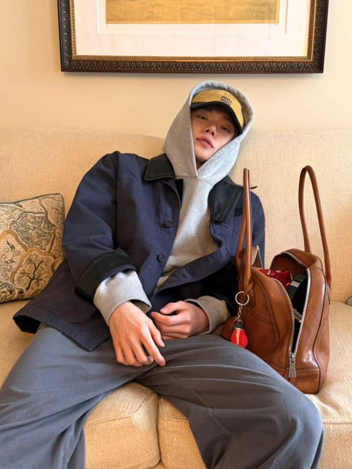 Yeonjun lounging on a cream-colored couch, wearing a gray hoodie and a navy jacket. They have a cap on and are relaxing with a brown handbag beside them.