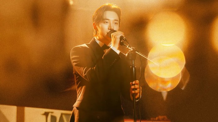 Lee Junho in a black suit performs on stage, holding a microphone under warm lighting with a blurred background.