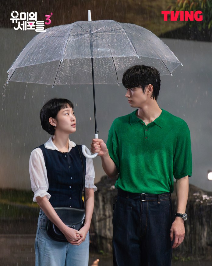 A scene featuringKim Go-eun and Kim Jae-won standing under a clear umbrella in the rain. Kim Go-eun, dressed in a navy outfit, looks up at Kim Jae-won, who is wearing a green shirt and holding the umbrella.