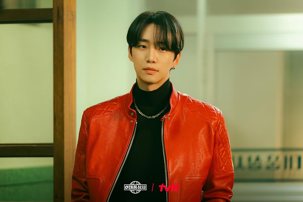 Lee Junho with dark hair wearing a bright red leather jacket over a black turtleneck sweater, standing against a softly lit background with wooden and green accents.